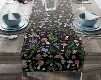 May include: Black table runner with a repeating pattern of mushrooms, ferns, flowers, and insects. The runner is on a wooden table with place settings.