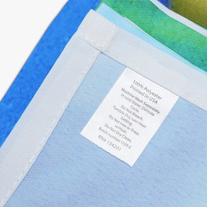 May include: A white care label with blue text detailing the care instructions for a 100% polyester item. The label reads "100% Polyester Printed in USA Machine Wash Separately in cold Water, Delicate Cycle. Do Not Bleach. Tumble Dry, Low Heat Setting. Do Not Iron or Press w/Heat. Do Not Dry Clean. Batch number 1220-C RN# 154201"