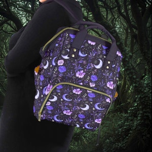 Può includere: Uno zaino viola e nero con un motivo floreale e lunare. Lo zaino ha più tasche e una chiusura a zip.