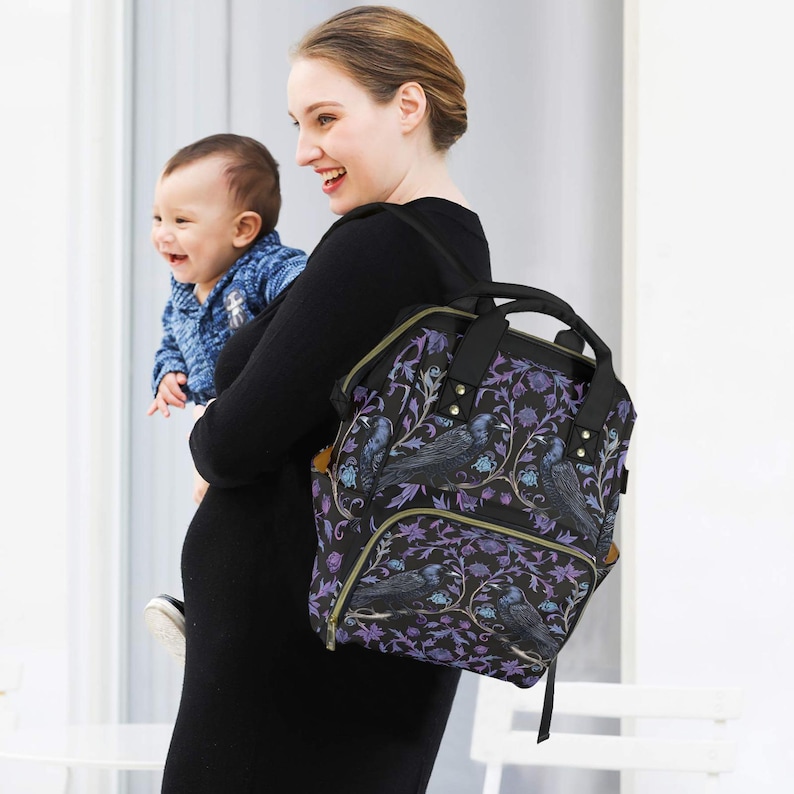May include: A black diaper bag with a floral and bird print. The bag has multiple pockets and a zipper closure. The bag is being worn by a person holding a baby.