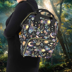Puede incluir: Mochila negra con diseño floral y celestial. El patrón incluye soles amarillos, lunas crecientes y varias flores en tonos azules, morados y amarillos. La mochila tiene un asa superior, un bolsillo frontal y cremalleras doradas.