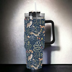May include: A stainless steel travel mug with a black handle and lid. The mug features a colorful pattern of owls, rabbits, snakes, mushrooms, and flowers on a dark blue background.