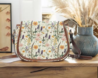 Könnte beinhalten: Eine braune Ledertasche mit einem cremefarbenen Canvas-Körper, der mit einem floralen Druck aus gepressten Wildblumen verziert ist. Die Tasche hat einen langen, verstellbaren Riemen.