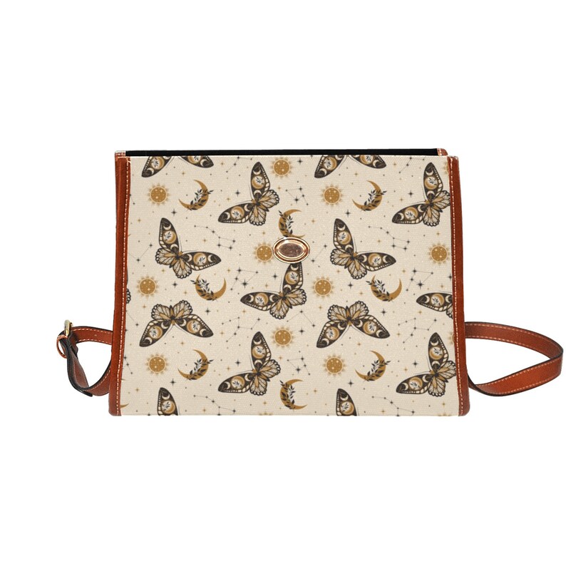 May include: A beige canvas crossbody bag with a brown leather strap and a gold metal clasp. The bag features a repeating pattern of black butterflies, moons, and suns.