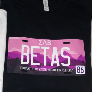 Op de afbeelding: Twee witte en zwarte T-shirts met een roze kentekenplaatontwerp. Het witte T-shirt heeft de tekst "ΣΛΓ GAMMAS" en "CULTURE IS PRIDE, PRIDE IS SUCCESS" met het nummer "90". Het zwarte T-shirt heeft de tekst "ΣΛΒ BETAS" en "OPPORTUNITY FOR WISDOM, WISDOM FOR CULTURE" met het nummer "86".
