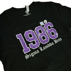 May include: Black t-shirt featuring a purple bandana print of the year 1986 and the text "Sigma Lambda Beta" in white.