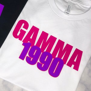 May include: Two t-shirts, one black and one white, with the words "BETA 1986" and "GAMMA 1990" printed on them in purple and white lettering.