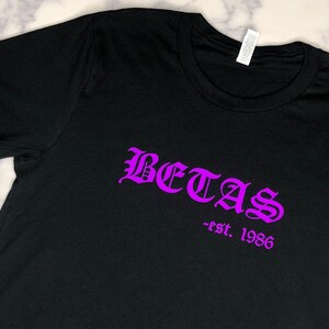Könnte beinhalten: Schwarzes T-Shirt mit einem violetten Aufdruck "BETAS - est. 1986".