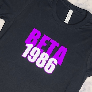 Könnte beinhalten: Schwarzes T-Shirt mit einem lila und weißen Aufdruck, der "BETA 1986" lautet.