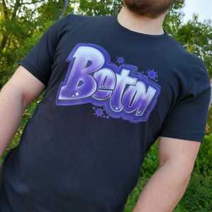 Può includere: Una maglietta nera con un design in stile graffiti viola e bianco che dice "Bety".