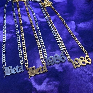 May include: Four necklaces with silver and gold chains. Two necklaces say "Beta" in a gothic font, and two say "1986". The necklaces are displayed on a purple fabric background.