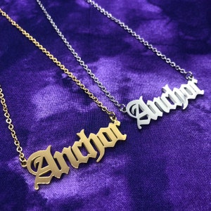 May include: Two gold necklaces with the word "Anchor" spelled out in a gothic font. The necklaces are on a purple background.