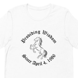 Puede incluir: Camiseta blanca con un gráfico negro de un caballo encabritado con el texto "Providing Wisdom Since April 4, 1986".