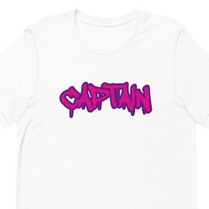 May include: White t-shirt with a pink and purple graffiti-style word "CAPTAIN" printed on the front.