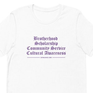Puede incluir: Camiseta blanca con el texto "Brotherhood Scholarship Community Service Cultural Awareness Established 1986" impreso en morado.