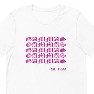 Puede incluir: Camiseta blanca con cuello redondo. La parte delantera presenta la palabra "GAMMAS" repetida cinco veces en un patrón apilado, en un color púrpura degradado. Debajo del texto apilado está el texto "-est. 1990" en púrpura.