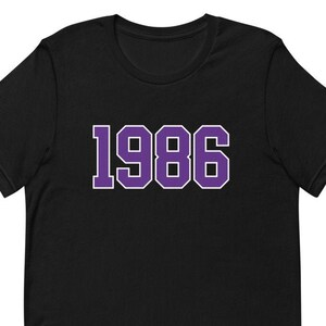 Könnte beinhalten: Schwarzes T-Shirt mit der Jahreszahl "1986" in lila Blockbuchstaben mit weißem Rand. Das T-Shirt hat einen Rundhalsausschnitt und kurze Ärmel. Der Stoff scheint weich zu sein.