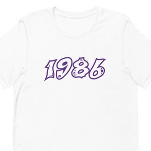 May include: White t-shirt with a purple 1986 graphic printed on the front.