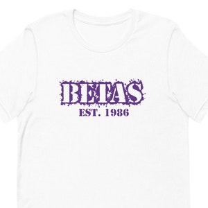 Könnte beinhalten: Weißes T-Shirt mit einem lila Aufdruck „BETAS“ über „EST. 1986“. Der Text hat ein verwaschenes, spritzerartiges Design. Das T-Shirt ist ein klassischer Rundhalsausschnitt.