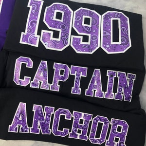 May include: Three black fabric patches with purple paisley designs and white outlines. The patches read "1990", "CAPTAIN", and "ANCHOR".