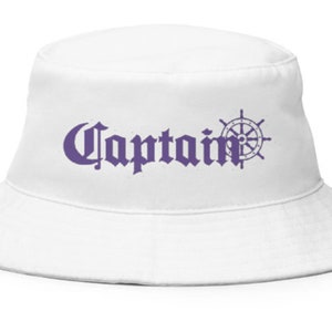 Puede incluir: Sombrero de pescador blanco con la palabra "Captain" en morado, en fuente gótica, acompañado de un timón morado. El sombrero tiene un ala ancha y una corona redonda, para proteger del sol.