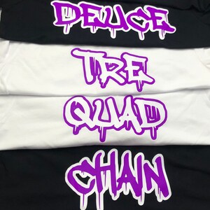 Könnte beinhalten: Ein Stapel weißer und schwarzer T-Shirts mit lilafarbenen Text. Der Text auf den T-Shirts lautet "Solo", "Captain", "Deuce", "Tre", "Quad", "Chain" und "Anchor".