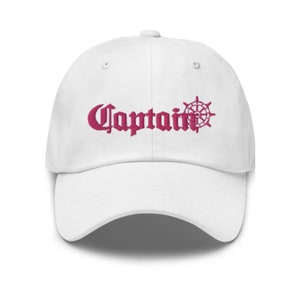 May include: A white baseball cap with the word "Captain" embroidered in a burgundy gothic font, accompanied by a ship's wheel design. The cap has a curved brim and an adjustable strap.