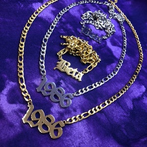 Könnte beinhalten: Drei goldene Ketten mit Anhängern. Die oberste Kette hat einen silbernen Anhänger mit der Aufschrift "Beta". Die mittlere Kette hat einen silbernen Anhänger mit der Aufschrift "1986". Die unterste Kette hat einen goldenen Anhänger mit der Aufschrift "1986".