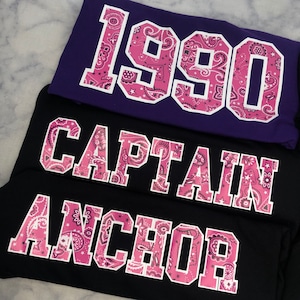 May include: Three black and purple sweatshirts with pink bandana print lettering. The top sweatshirt says "1990", the middle sweatshirt says "CAPTAIN", and the bottom sweatshirt says "ANCHOR".