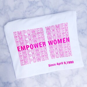 May include: White t-shirt with the phrase "EMPOWER WOMEN" repeated in pink lettering. The text is stacked vertically and the phrase "Since April 9, 1990" is printed below the stacked text.