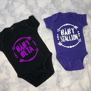May include: Two baby onesies, one black with purple text that says "Baby Beta" and one purple with white text that says "Baby Stallion".