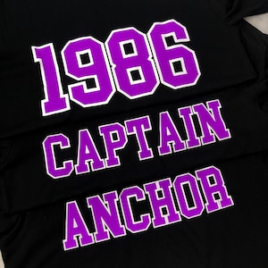 Könnte beinhalten: Schwarzes T-Shirt mit violetter Aufschrift "1986", "CAPTAIN" und "ANCHOR".