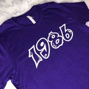 Könnte beinhalten: Lilafarbenes T-Shirt mit weißem Text, der in einer stilisierten Schrift "1986" lautet.