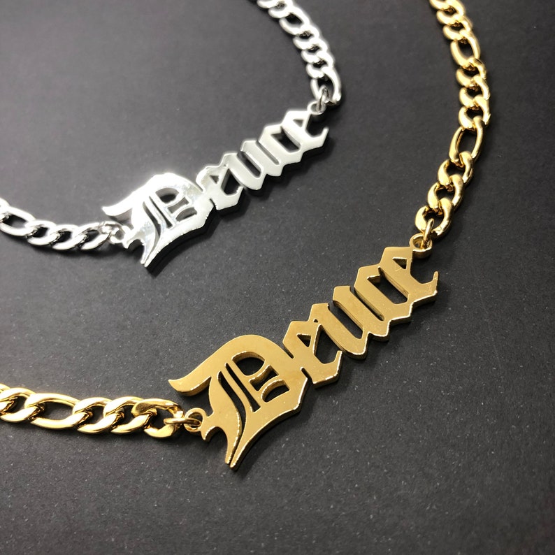 K&ouml;nnte beinhalten: Zwei goldene Ketten mit dem Wort "Peace" in altenglischen Lettern. Die obere Kette ist silber und die untere Kette ist gold.