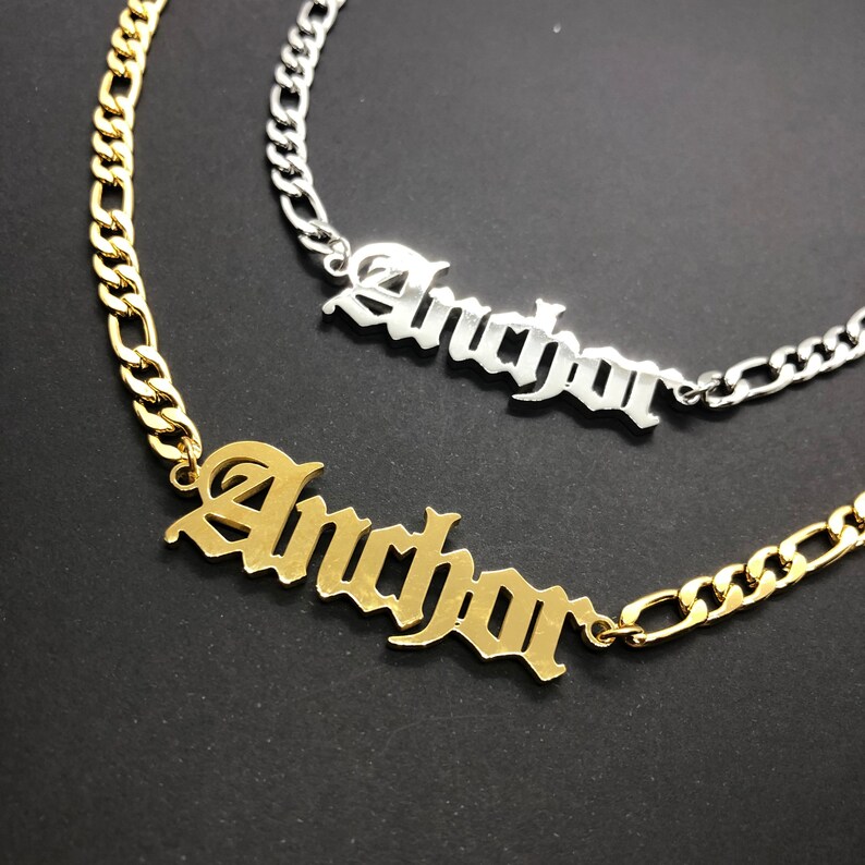 K&ouml;nnte beinhalten: Zwei goldene und silberne Halsketten mit einem kursiv geschriebenen 'Anchor'-Anh&auml;nger. Die Halsketten liegen auf einem schwarzen Hintergrund.