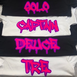 May include: Four white t-shirts with different names written in pink and purple graffiti style. The names are "Solo", "Captain", "Deuce", and "Tre".