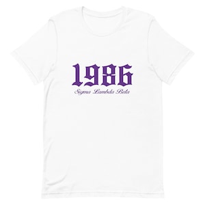Op de afbeelding: Wit T-shirt met een paarse 1986 in gotische letter en de tekst "Sigma Lambda Beta" eronder.