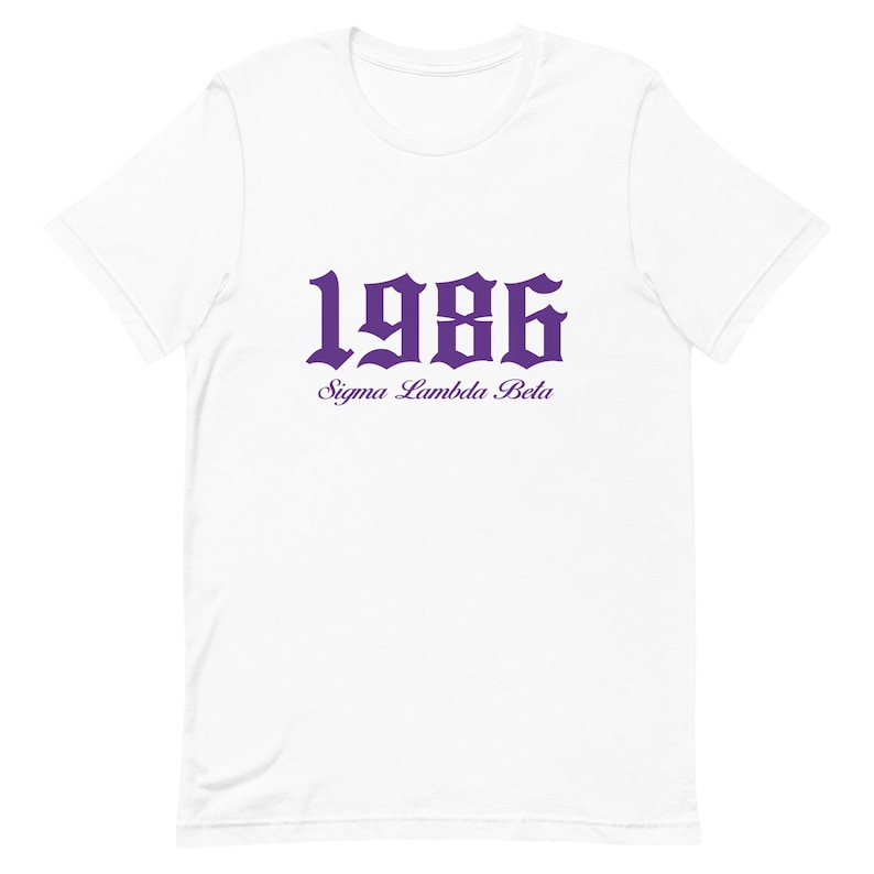 Op de afbeelding: Wit T-shirt met een paarse 1986 in gotische letter en de tekst "Sigma Lambda Beta" eronder.