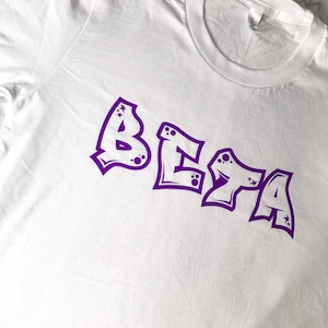 May include: White t-shirt with the word "BETA" written in a purple graffiti style font. The letters have small stars and dots around them.