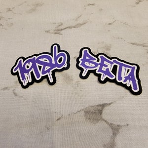 May include: Two purple and white stickers with black outlines. The stickers have the words "1986" and "BETA" written in a graffiti style.