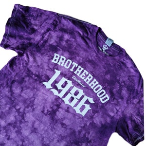 Op de afbeelding: Paarse tie-dye T-shirt met witte tekst die "Brotherhood Established 1986" zegt.