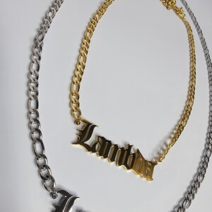 May include: A gold chain necklace with a nameplate pendant that reads "Lambda" in a gothic font. The chain is a curb style and is paired with a silver chain.