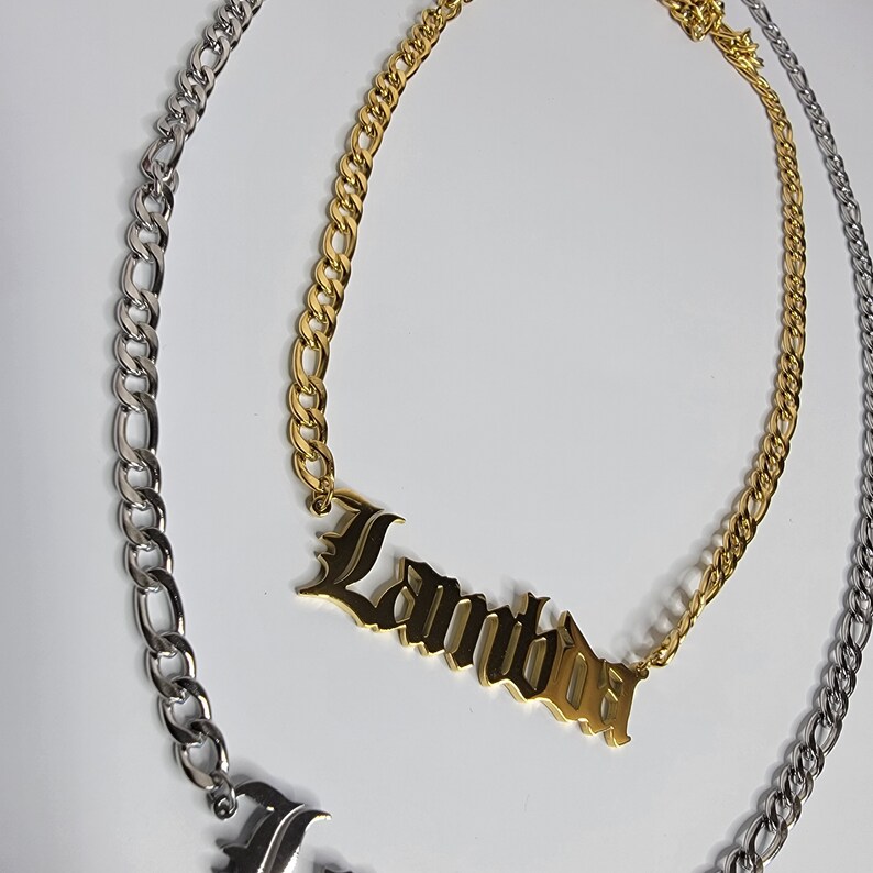 May include: A gold chain necklace with a nameplate pendant that reads "Lambda" in a gothic font. The chain is a curb style and is paired with a silver chain.