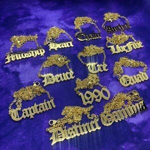 May include: A collection of gold necklaces with different names and words in a gothic font. The names include "Friendship", "Sean", "Chain", "Anchor", "Live Free", "Deuce", "Tre", "Quad", "1990", "Gamma", and "Distinct".