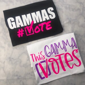 May include: Two white t-shirts with black and purple text. The first shirt says "GAMMAS #VOTE" in black and pink text. The second shirt says "This GAMMA Votes" in purple and pink text.