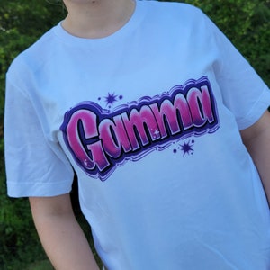 May include: White t-shirt with a pink and purple graffiti-style design that says "Gamma" with white stars.