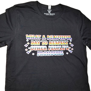 May include: Black t-shirt with the text "What a beautiful day to respect other people's pronouns" in rainbow colours.