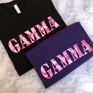 Op de afbeelding: Twee T-shirts met het woord "GAMMA" bedrukt in roze camouflage letters. Een shirt is zwart en de andere is paars.