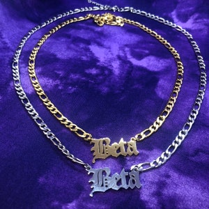 Könnte beinhalten: Zwei goldene und silberne Kettenhalsbänder mit dem Wort "Beta" in gotischer Schrift auf jedem Anhänger.