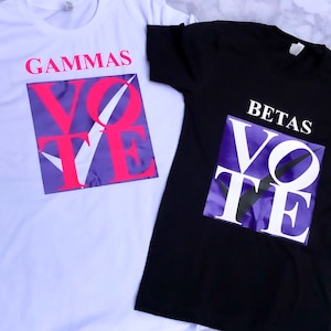 May include: Two t-shirts with the word "VOTE" printed on them. The shirt on the left is white with pink letters and a purple background. The shirt on the right is black with white letters and a purple background. The text "GAMMAS" is printed above the word "VOTE" on the white shirt. The text "BETAS" is printed above the word "VOTE" on the black shirt.